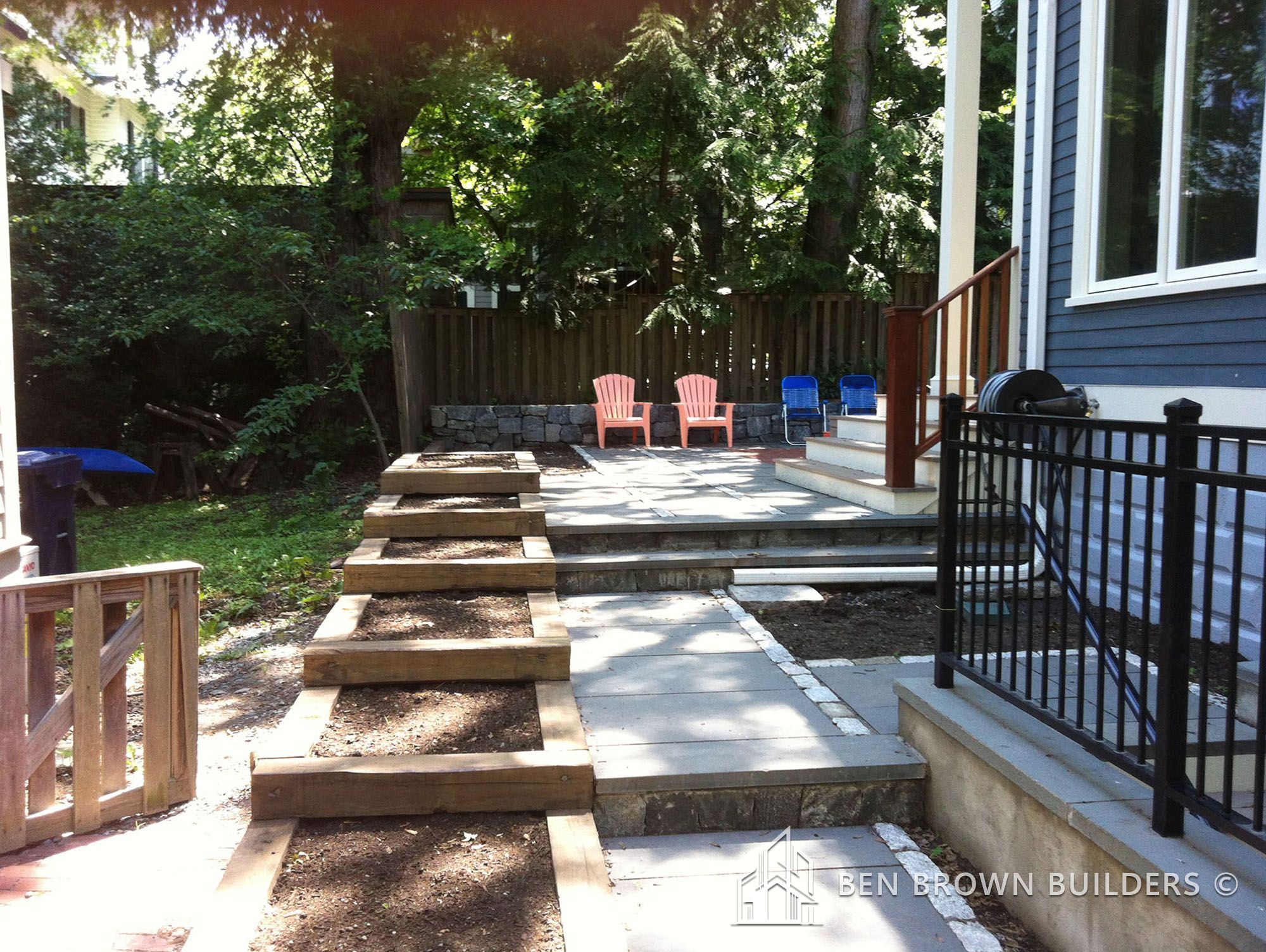 Backyard patio with wooden steps leading to a seating area with two red Adirondack chairs and a fenced garden