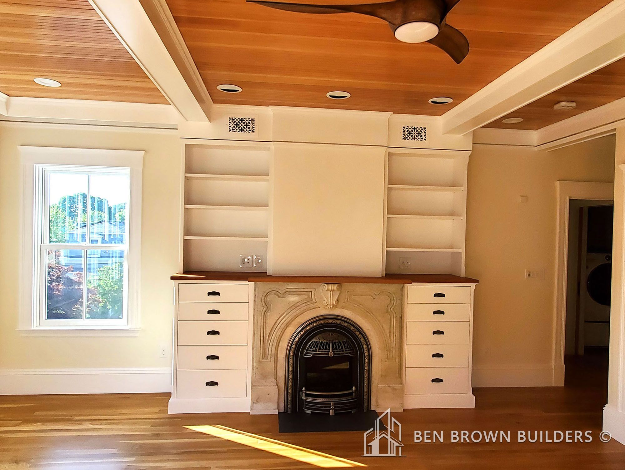 Bright room with a white fireplace, built-in shelving, hardwood floors, and a wooden ceiling