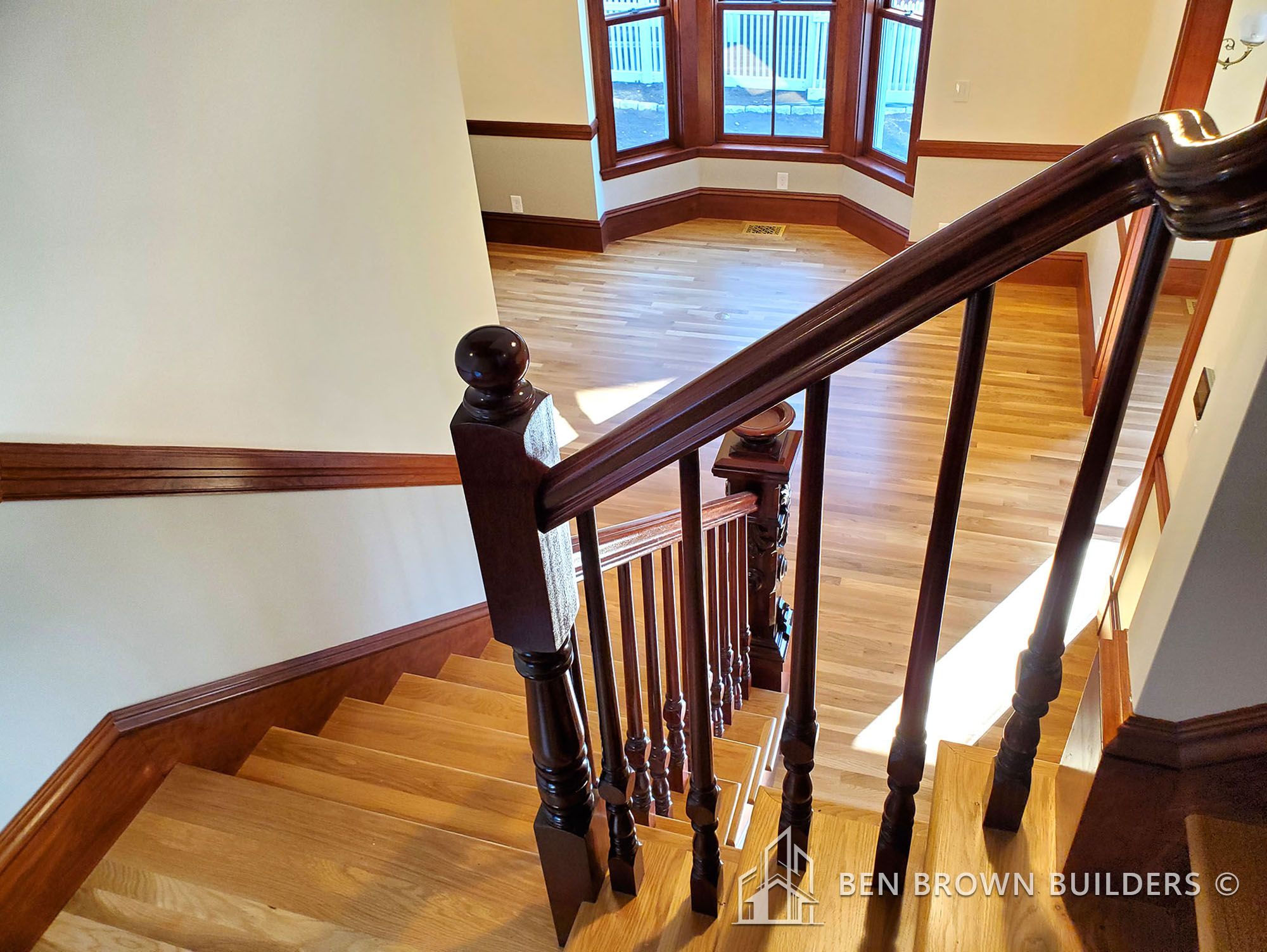 View from the top of a wooden staircase with ornate handrails, overlooking a sunlit room with bay windows